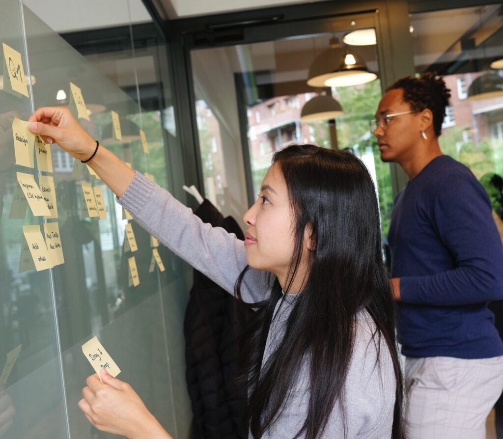 Two people adding notes to a wall for roadmap planning. One person is a young woman with long black hair. The second person is a has their hair pulled back with glasses.