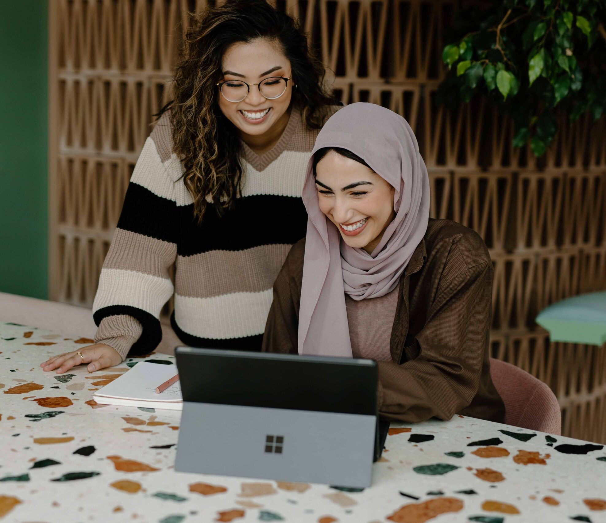 two women laughing together at a computer. one woman has curly brown hair and is wearing glasses and a stripped sweater. The second woman is wearing a tan hijab and brown jacket.