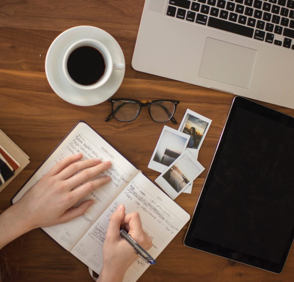 overhead view of a workspace. A computer, glasses, coffee cup, photos, and the hands of someone writing in a a notebook